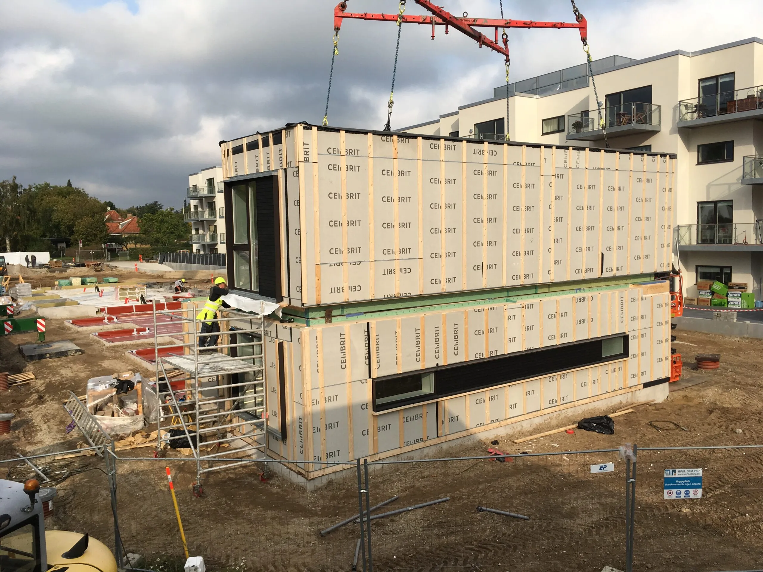 Kran løfter præfabrikeret boligmodul med Cembrit-beklædning på byggepladsen, færdige lejligheder i baggrunden